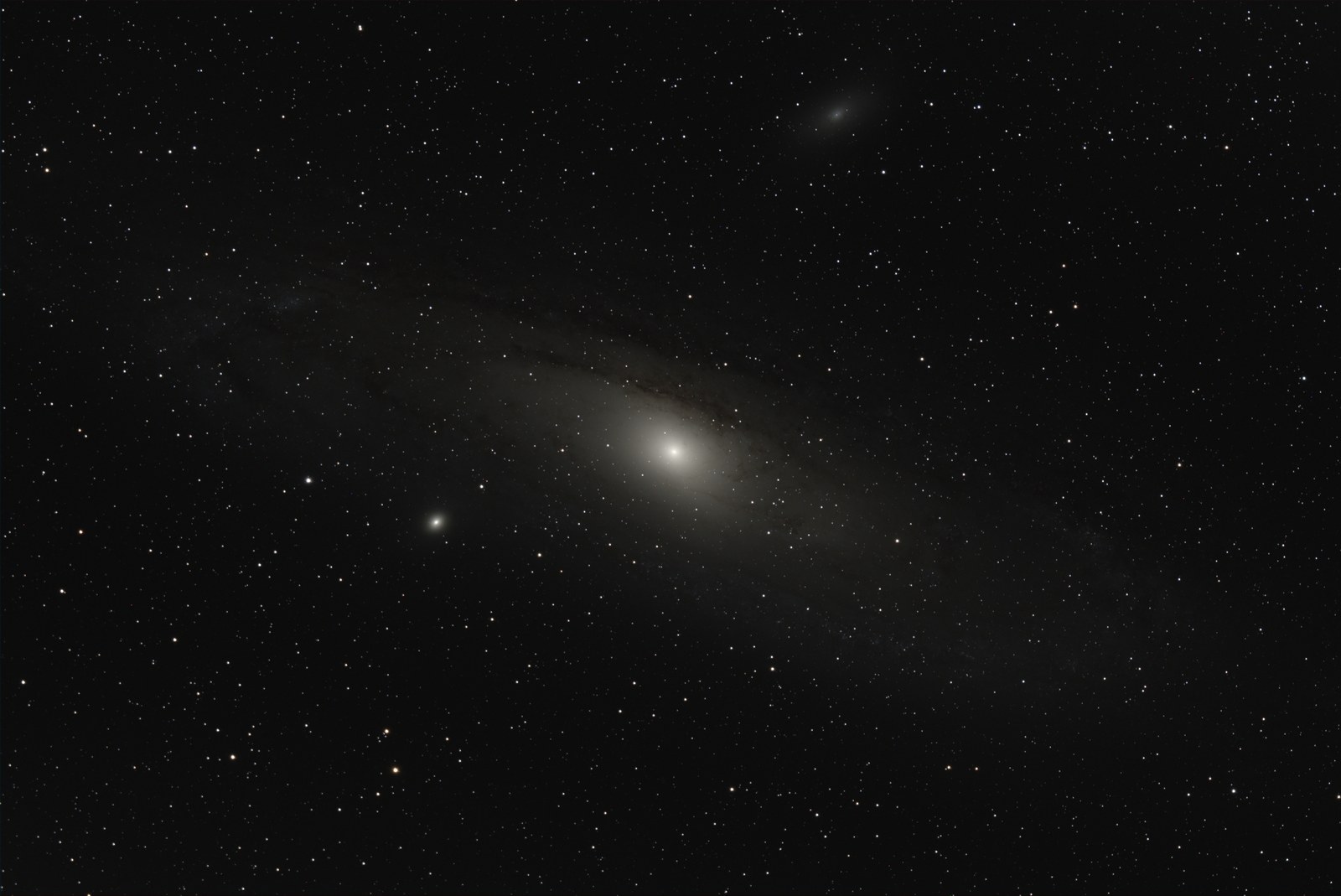 The Andromeda galaxy stretched across the whole frame at a tilt, a bright core fading into a wide disk with faint dust lanes, the small round companion M32 just below the core and the fainter oval M110 above, in a field of stars.