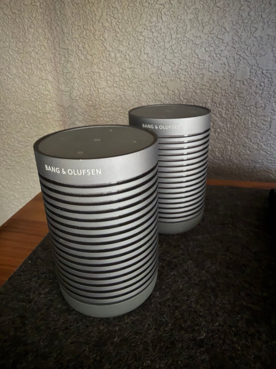 Two small silver Bang and Olufsen speakers, ribbed cylinders with dark tops, on a shelf against a textured wall.