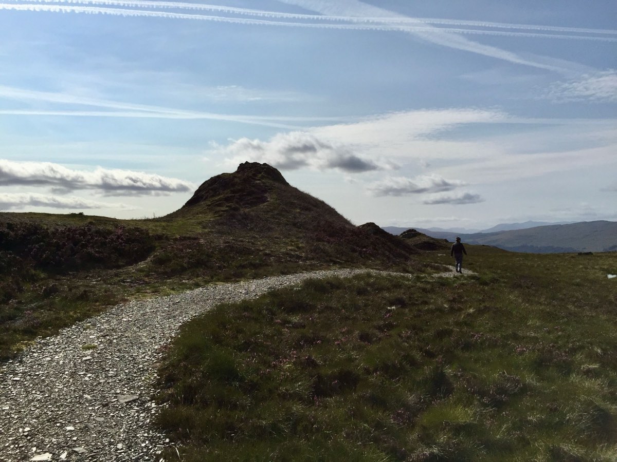 A man walking ahead on a gravel path toward a small rocky hill, contrails crossing the sky.